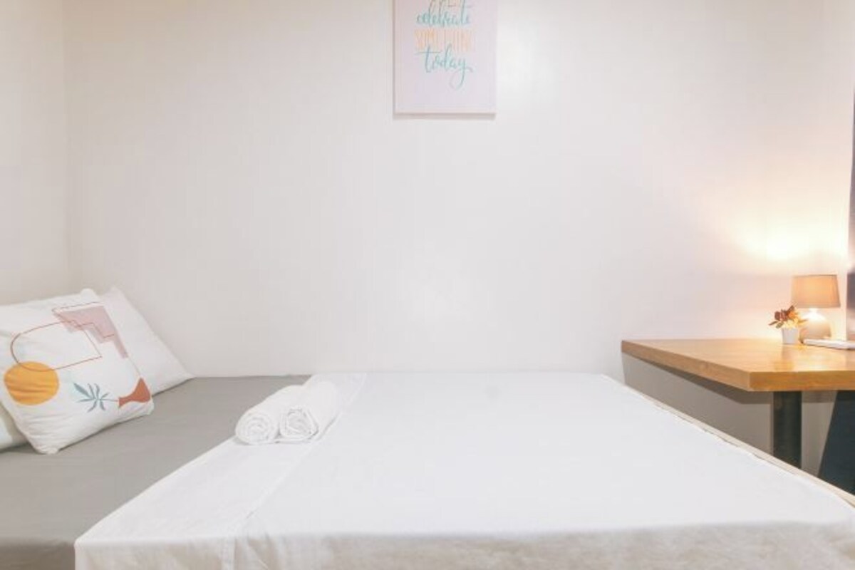 A single bed with a white bedspread, a bedside table with a lamp, and a wall-mounted picture frame.