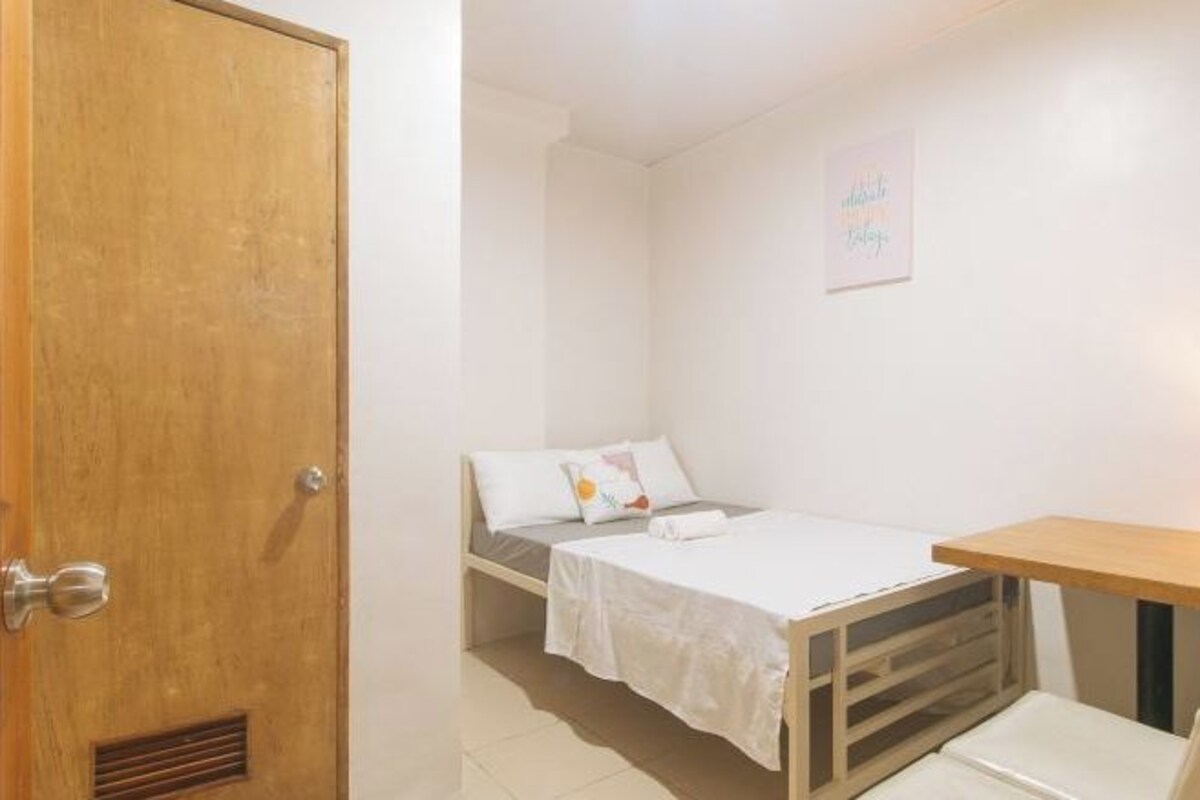 Una habitación pequeña y limpia con puerta de madera, una cama con ropa blanca y una mesita.
