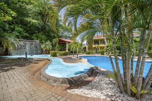 A swimming pool surrounded by palm trees and a stone wall. - Peaceful Condo, Pool View, Access to 7 common Pools, Gated, a minute drive beach (Playa Ocotal)