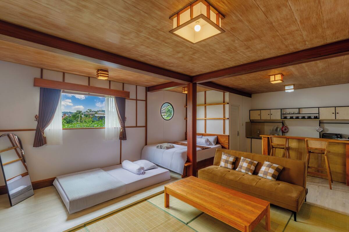 A traditional Japanese-style room with tatami flooring, a low wooden bed, a sofa, and a kitchen area with wooden cabinets and a bar stool.