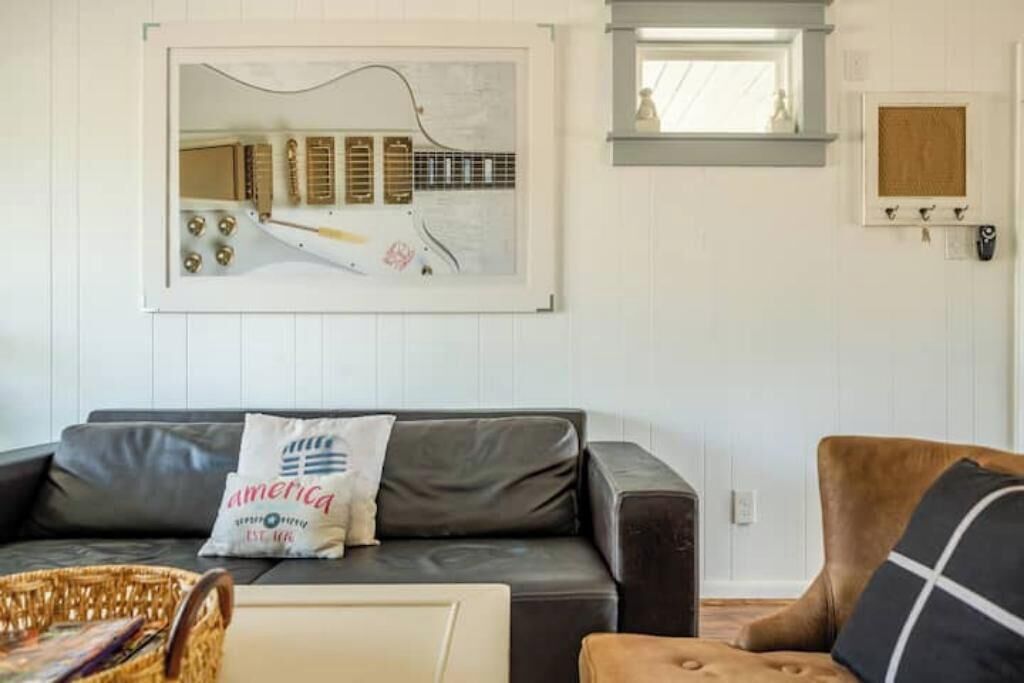 A living room with a grey sofa, a brown armchair, a framed abstract painting, and a white wall with a small window and a wooden cabinet.