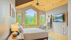 Un dormitorio con una cama grande, ventilador de techo y televisor. Se ven ventanas con vistas a árboles y montañas.