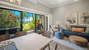 Una sala de estar moderna con un amplio ventanal corredero que da a vistas de un jardín tropical, un rincón de descanso acogedor con un sofá azul y un taburete estampado, y una mesa de comedor de madera.