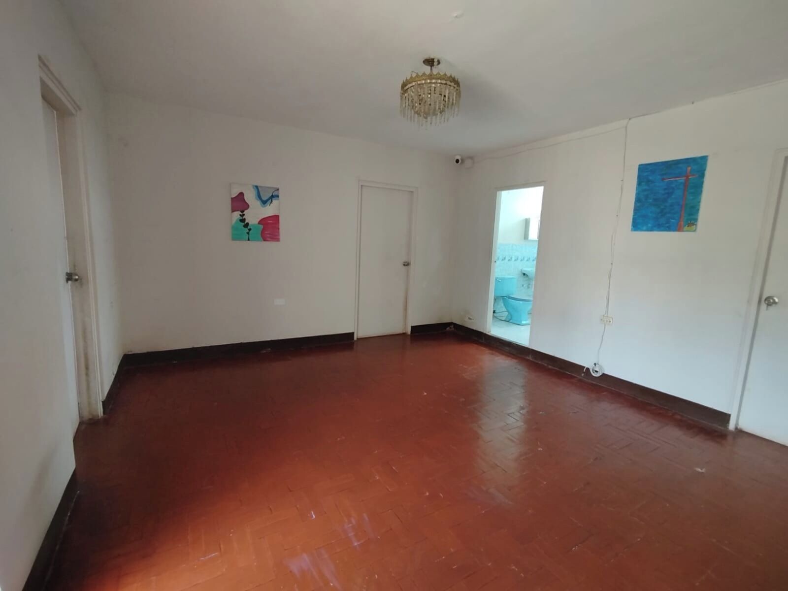 Una habitación amplia con suelo de madera, paredes blancas y una araña de cristal. Se observan dos puertas, una de ellas da acceso a un ba ño con inodoro a la vista.