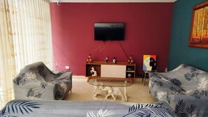 A room with a red wall, a mounted TV, a wooden cabinet with decorative items, and two patterned armchairs.