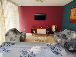Una habitación con una pared roja, un televisor montado, un mueble de madera con objetos decorativos y dos sillones estampados.