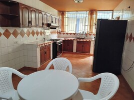 Una cocina con armarios de madera, una estufa y un refrigerador. Hay dos sillas de plástico blanco y una mesita pequeña.