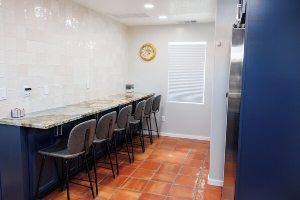 A kitchen with a marble countertop, four bar stools, a clock on the wall, and a refrigerator.