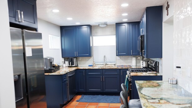A modern kitchen with dark blue cabinets, a stainless steel refrigerator, a tiled backsplash, and a large island with a granite countertop.