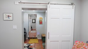 A room with a sliding door leading to a hallway, a bed with colorful bedding, and a small bench.