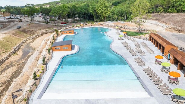 Une grande piscine à la forme incurvée, entourée de chaises longues et de parasols, avec en toile de fond des collines et des arbres.