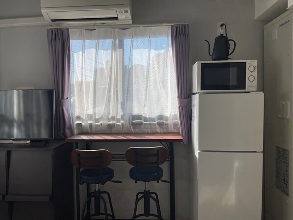 Cocina compacta con microondas, refrigerador y aire acondicionado sobre la ventana. Hay dos taburetes y una mesa pequeña frente a la ventana.
