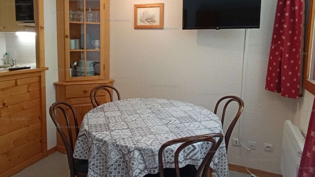 Un coin repas avec une table ronde, des chaises, un meuble avec des portes en verre, un téléviseur fixé au mur et un rideau rouge.