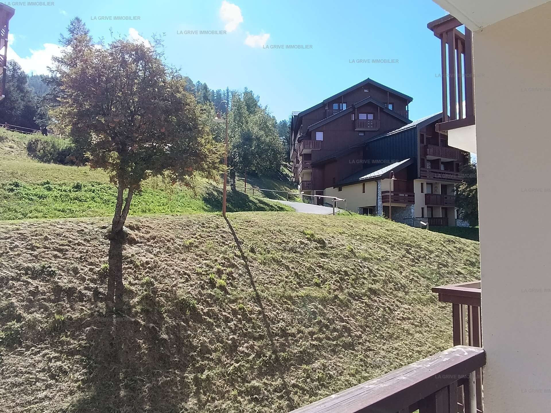 A residential building with a balcony, a tree in the foreground, and a hilly landscape in the background.