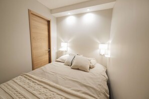 A bedroom with a wooden door, two wall-mounted lights, a bed with a white duvet and pillows, and a textured blanket. - Magnificent 3-room apartment 31m², south-east facing, mountain/resort view (VAL THORENS)