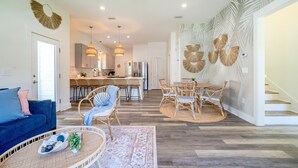 A spacious living area with a blue sofa, wicker chairs, a round table, and a kitchen in the background. - Walk2Beach•Free Golf Cart•YOLO Bikes (Santa Rosa Beach)
