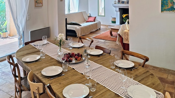 A dining area with a wooden table set for six, surrounded by wooden chairs. A large woven umbrella light fixture hangs above the table. - Au coeur des ocres - Charming vacation home (Roussillon)