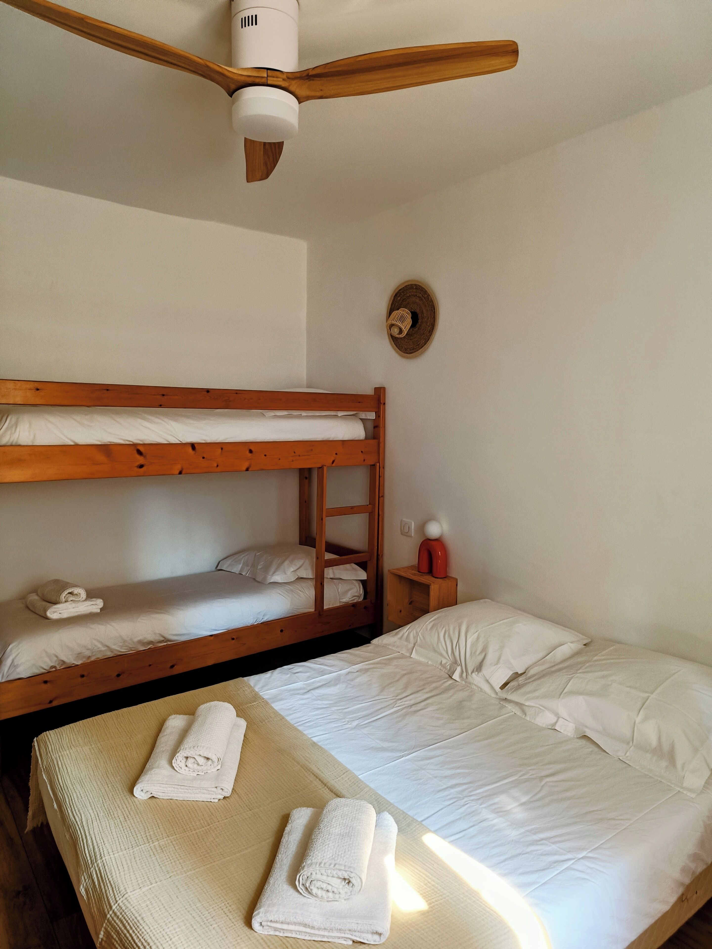 Une chambre individuelle avec un lit superposé, un lit simple, un ventilateur de plafond, une lampe fixée au mur et une petite table de chevet sur laquelle se trouve un objet rouge.