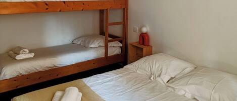 Une chambre individuelle avec un lit superposé, un lit simple, un ventilateur de plafond, une lampe fixée au mur et une petite table de chevet sur laquelle se trouve un objet rouge.