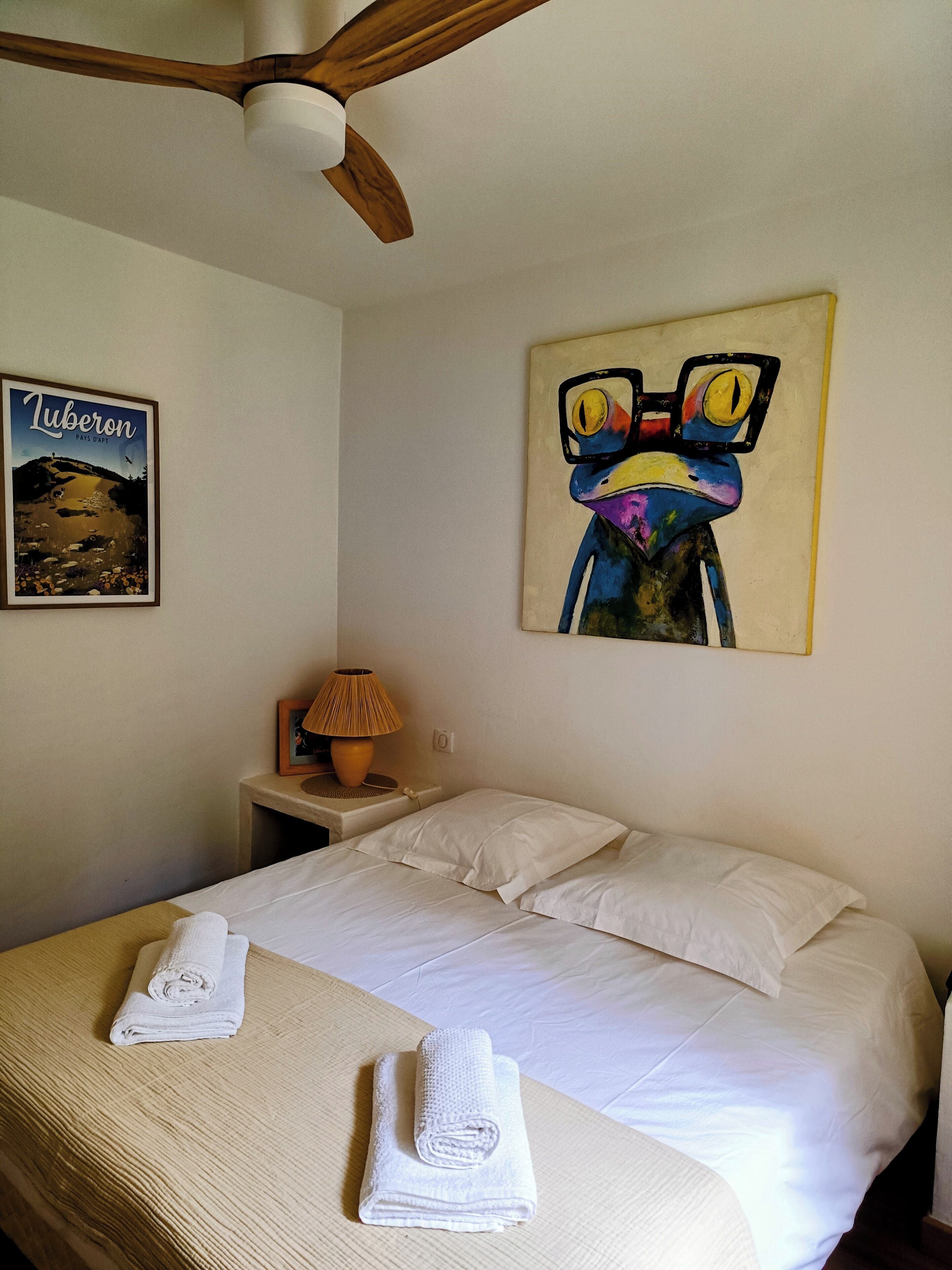 A hotel room with a bed, a ceiling fan, a painting, a lamp, and a framed picture on the wall.