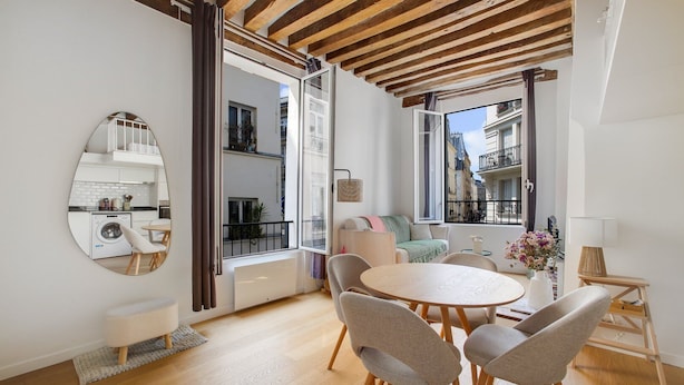 A modern living room with a round table, four chairs, a sofa, a large mirror, and a view of the city through large windows.