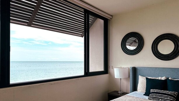 A bedroom with a large window, a bed with patterned bedding, a nightstand, and two wall-mounted mirrors. - Golden Hour 2BR Ocean View Apt (Holbox)