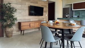 A modern dining area with a round wooden table, grey chairs, a flat-screen TV mounted on a wooden wall, and a kitchen area with a built-in oven and sink. - Golden Hour 2BR Ocean View Apt (Holbox)