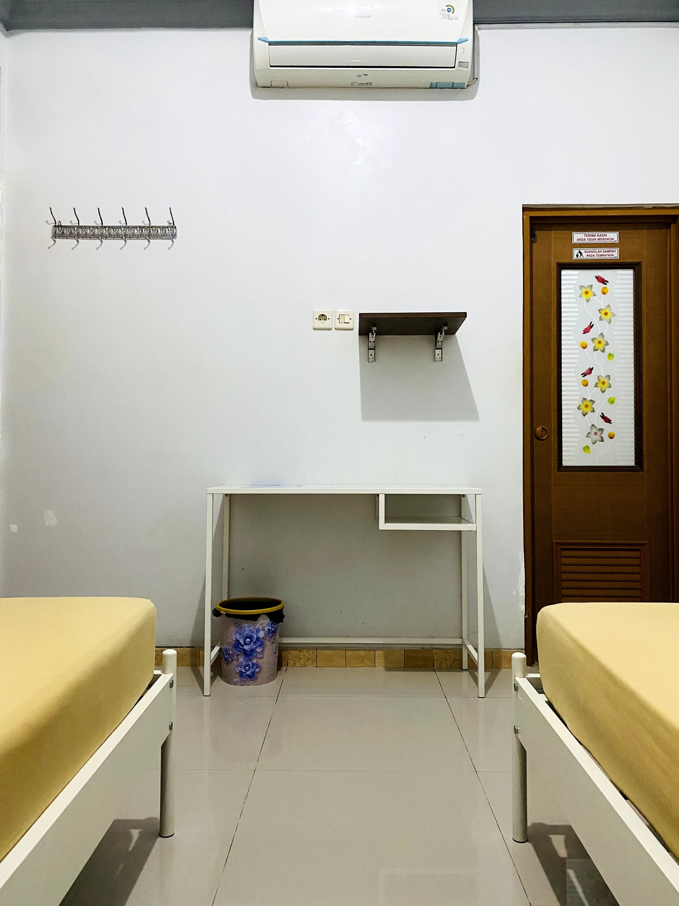 Una stanza con due letti, una scrivania, l'aria condizionata, un cestino e una porta con un adesivo decorativo.