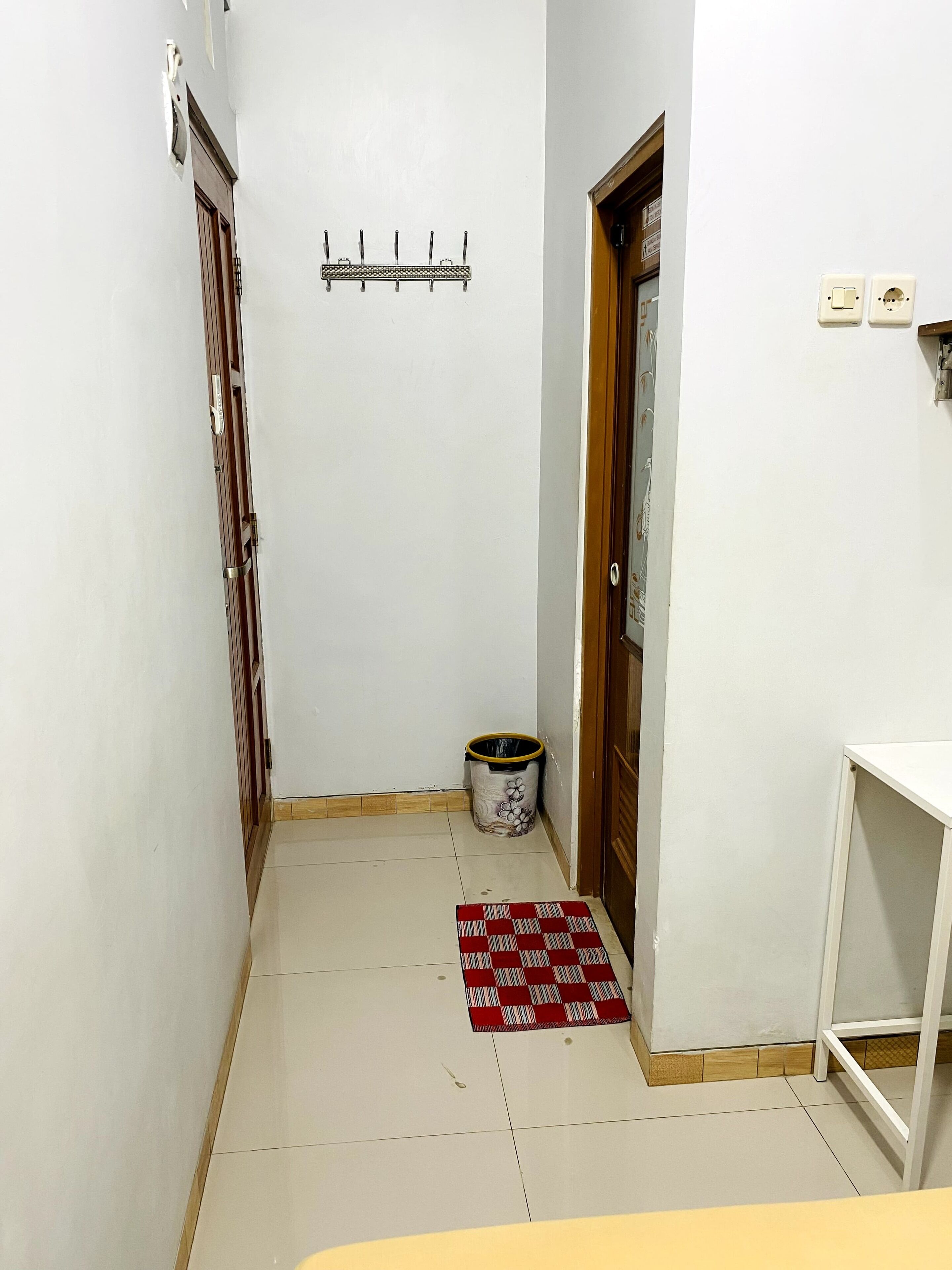 Un pasillo estrecho con piso de baldosas, una puerta de madera, un pequeño bote de basura y un tapete blanco y rojo a cuadros.