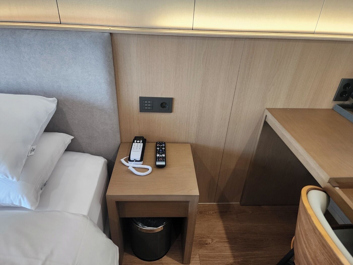 A compact room with a bed, a small bedside table with a phone and remote, a trash bin, and a wooden desk.