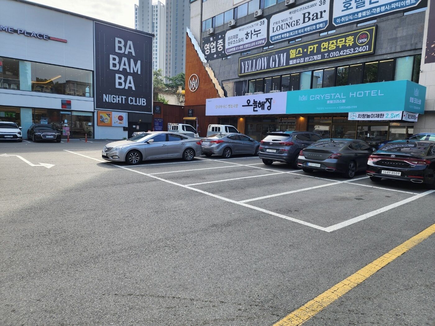 Un parking avec plusieurs voitures, un bâtiment portant une enseigne indiquant « BA BAM BA NIGHT CLUB », et une autre enseigne indiquant « YELLOW GYM ».