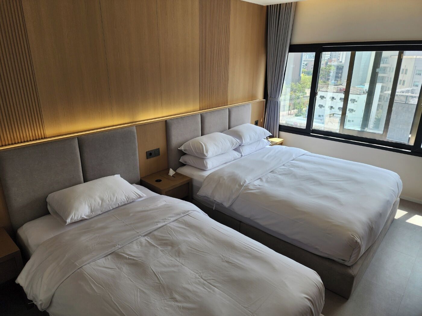 Habitación de hotel con dos camas, cabecera de madera, mesita de noche con lámpara y ventana con vista a la ciudad.