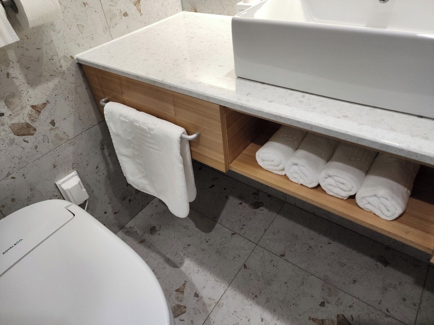 Ein Badezimmer mit einer weißen Toilette, einem Handtuch auf einem Haken und einem Regal mit ordentlich gefalteten Handtüchern.