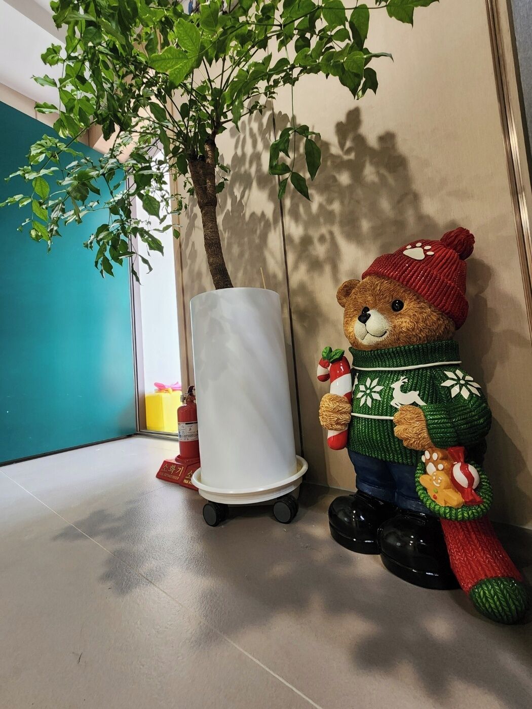 A teddy bear wearing a sweater and hat, a white cylindrical object, and a tree with green leaves.