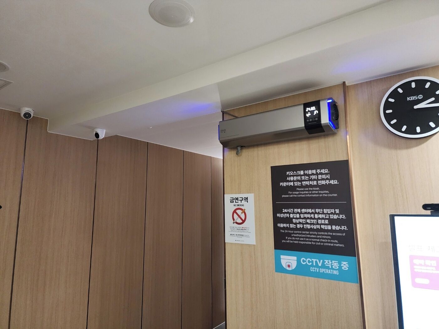 A room with a wooden panel wall, a clock, a CCTV camera, and a no-smoking sign.