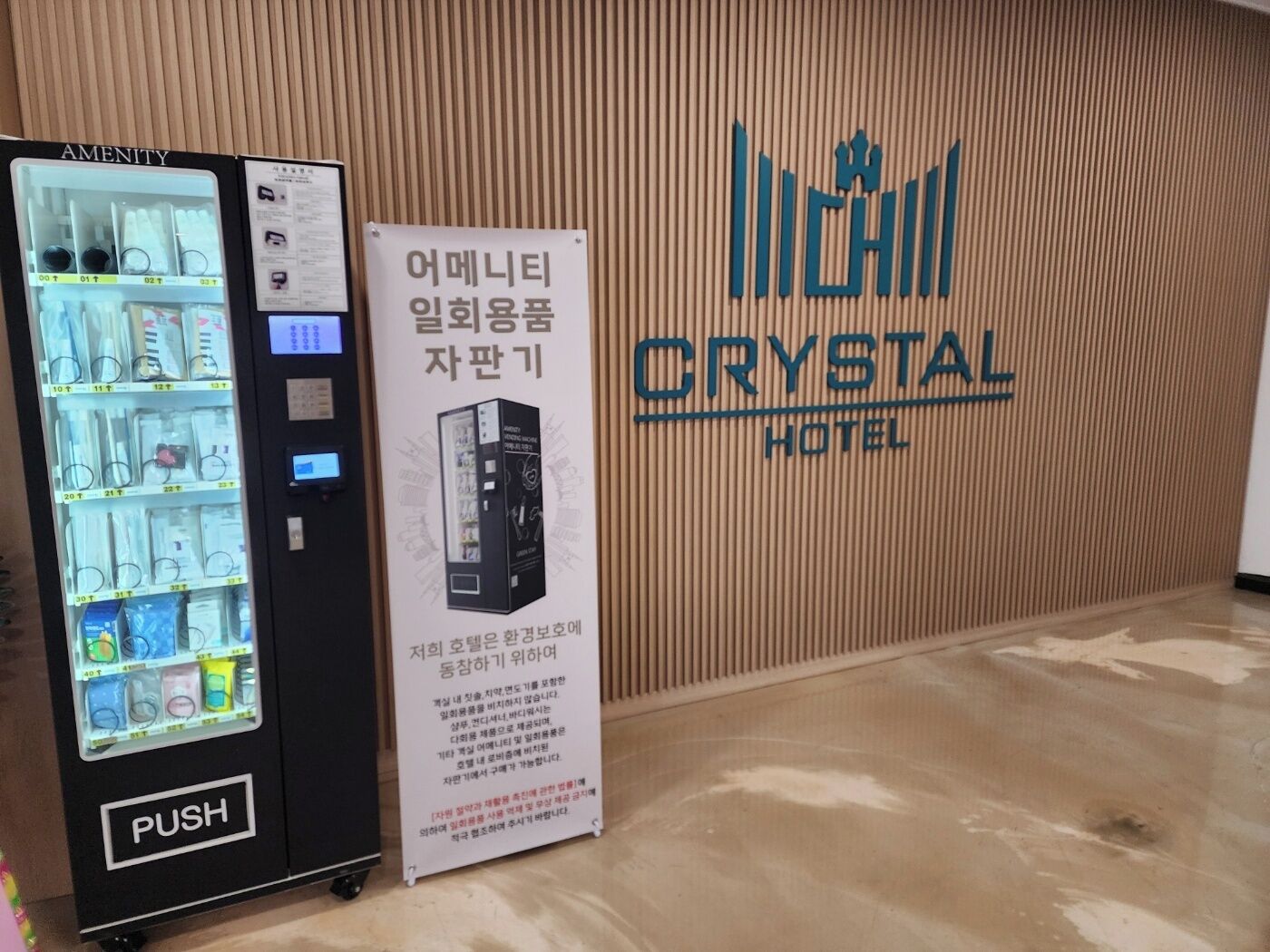 Una máquina expendedora con un botón de 'PUSH', un letrero con texto coreano y el rótulo del 'Crystal Hotel' en una pared de madera.