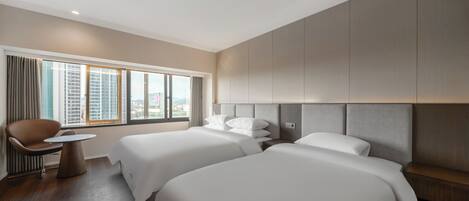 Habitación de hotel con dos camas, una silla, una mesita y un amplio ventanal con vistas a la ciudad.