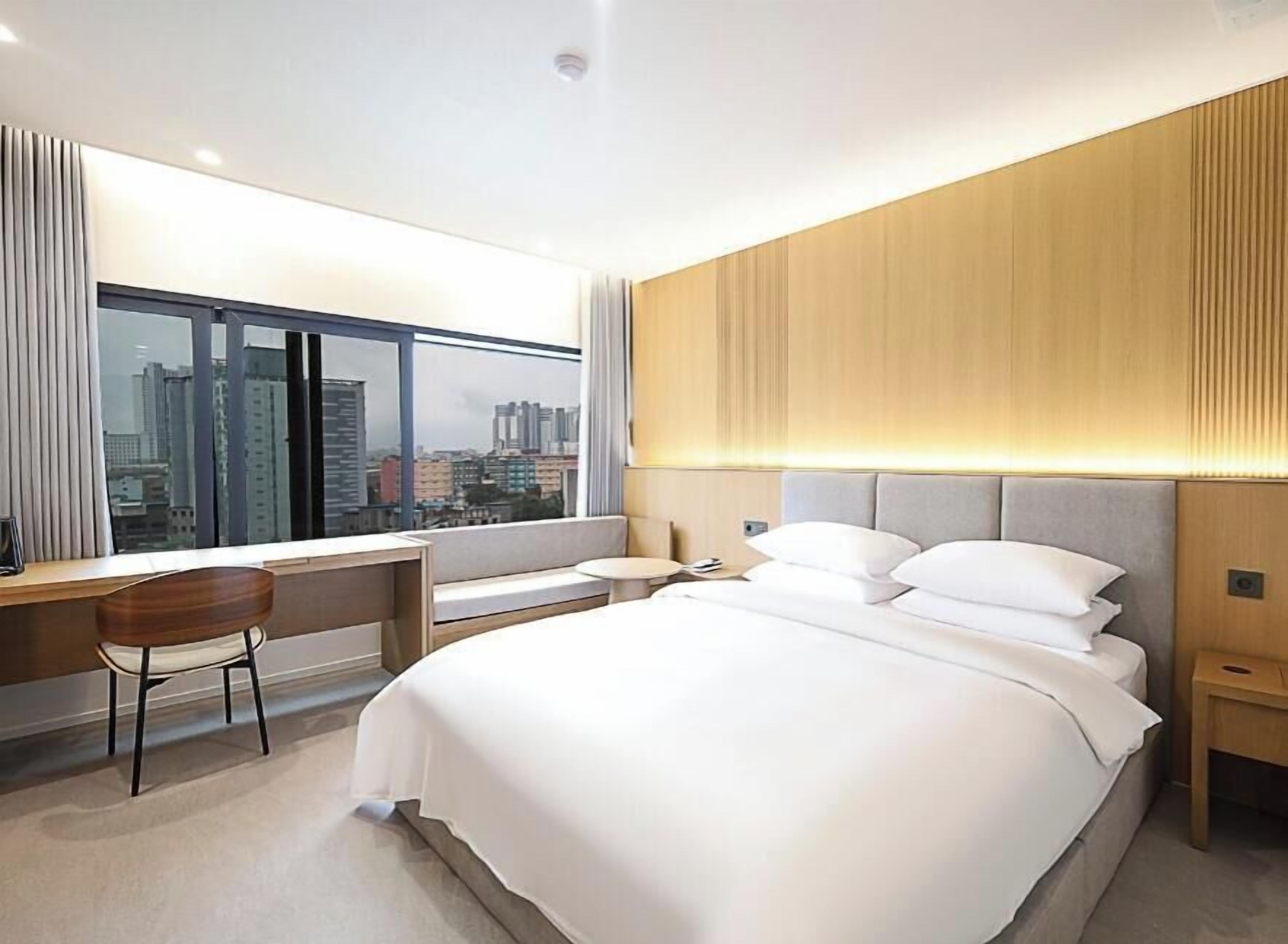 A modern hotel room with a large bed, a desk with a chair, a bedside table, and a view of the city through large windows.