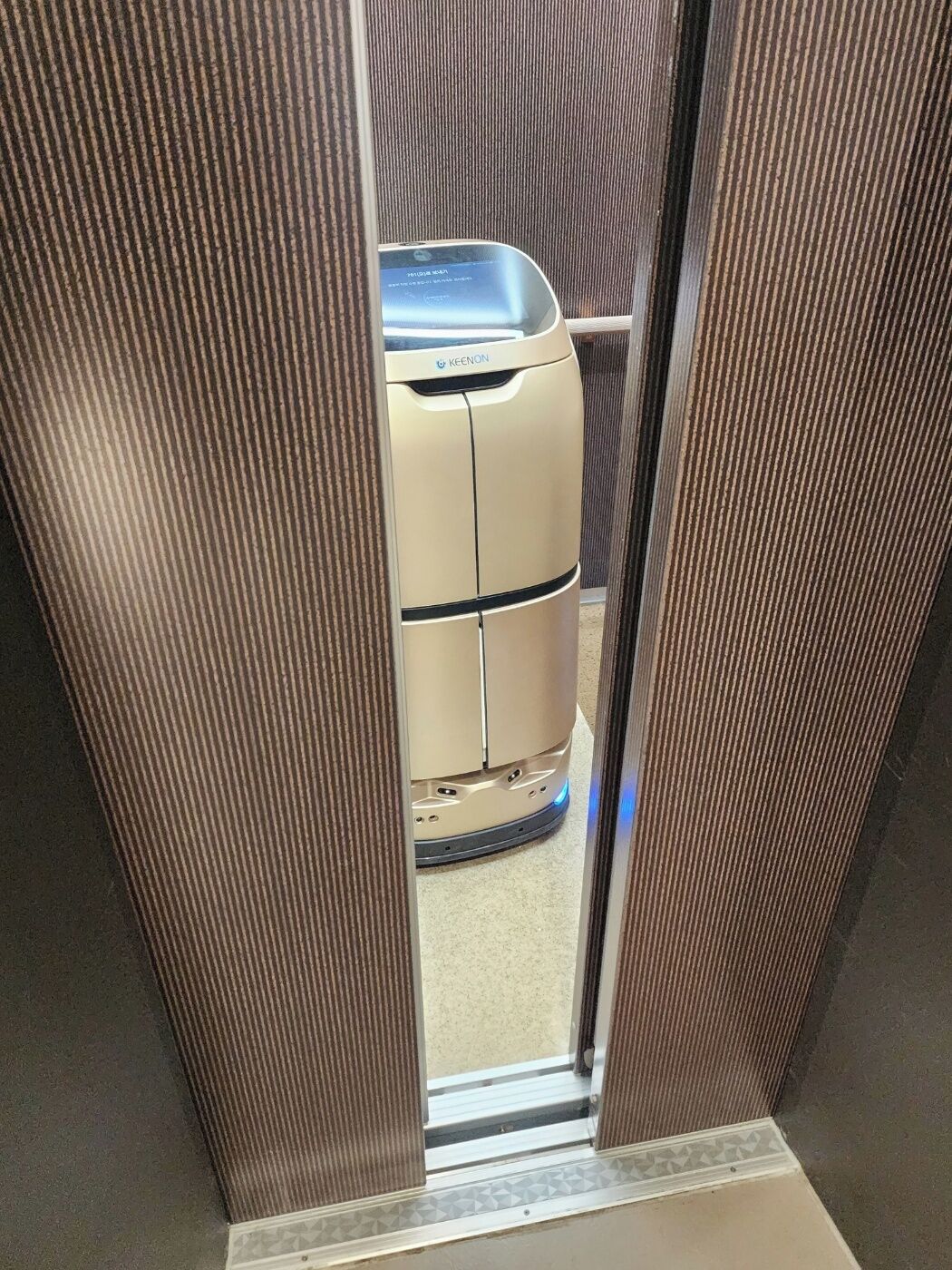 Une voiture beige, de petite taille et à deux portes, est garée à l’intérieur d’une pièce dont un mur est orné de motifs texturés en bandes verticales.