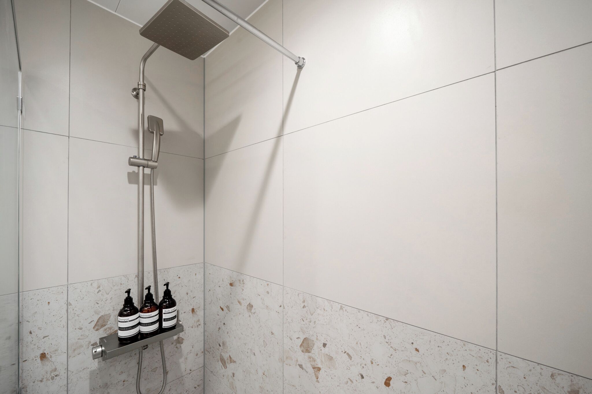 A shower with a handheld showerhead, a wall-mounted faucet, and three bottles of shampoo on a shelf.