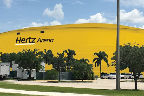 A large yellow building labeled "Hertz Arena" with trees and parked cars in the foreground.