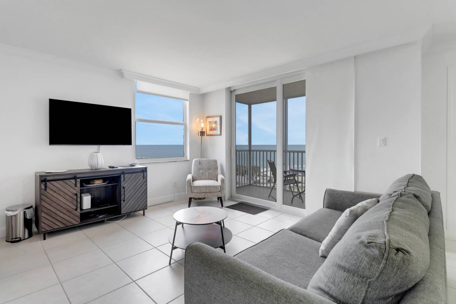 A modern living room with a grey sofa, a small round table, a TV mounted on the wall, and a balcony with a view of the sea.