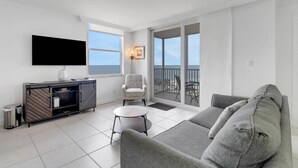 Una sala de estar moderna con un sofá gris, una pequeña mesa redonda, un televisor montado en la pared y un balcón con vista al mar.