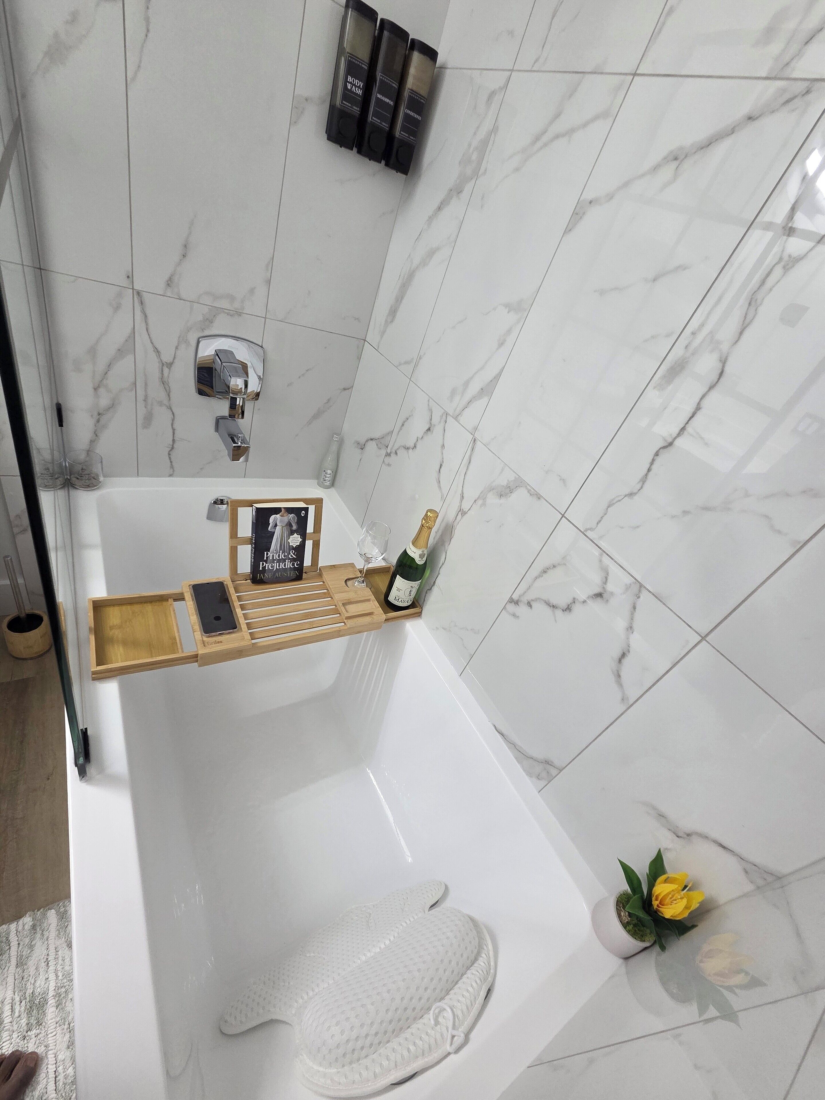 A modern bathroom with a marble-tiled wall, a wooden tray with a bottle of champagne, and a pair of slippers on the floor.
