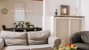 A modern living room with a beige sofa, a dark grey armchair, and a dining area with a table set for two.
