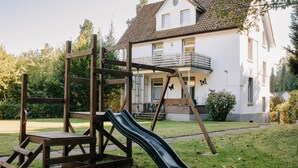A playground with a slide and climbing structure in front of a white house with a balcony.