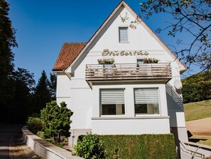 A white building with the name "Eibach" on the facade, a balcony with potted plants, and a clear blue sky in the background.