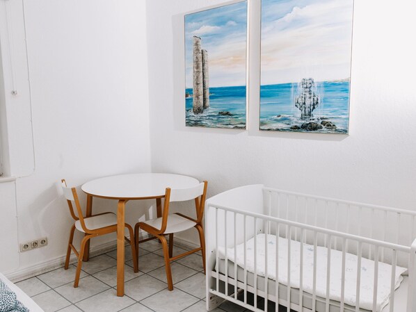 A room with a white crib, a round table with two chairs, and two paintings of coastal scenes on the wall.