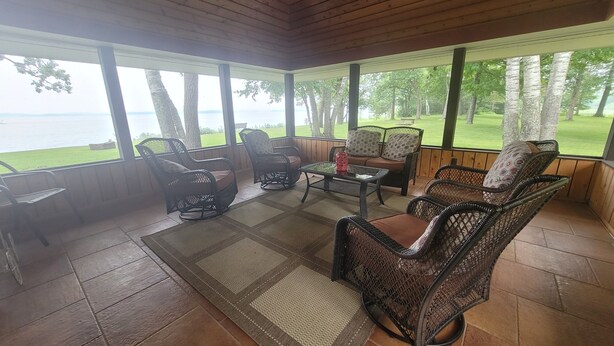 A covered patio with wicker furniture, a glass coffee table, and a view of a grassy area and water.