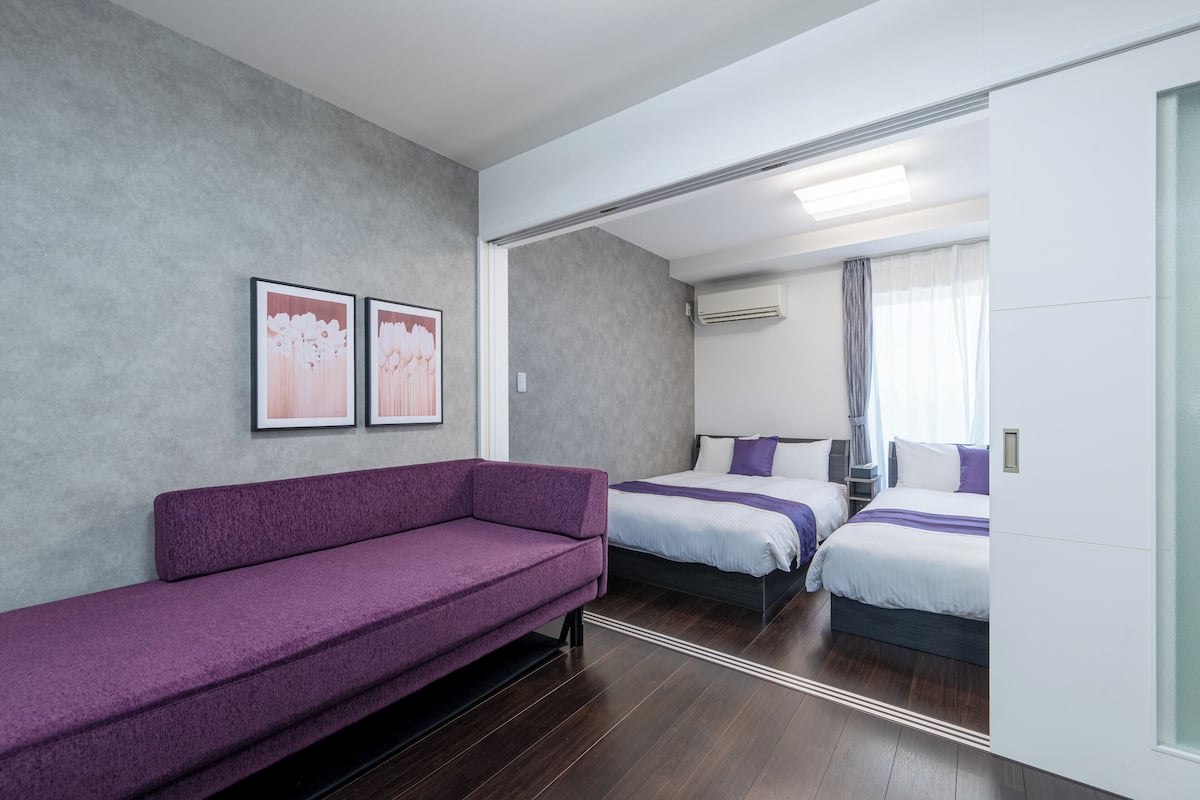 Une chambre d’hôtel moderne avec un canapé violet, deux lits avec une literie assortie, un mur d’accent gris et des œuvres d’art encadrées.